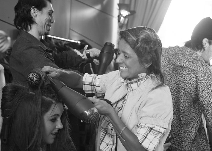 A PANYC stylist blow-dries a model's hair backstage, smiling as she works, shot in black and white.